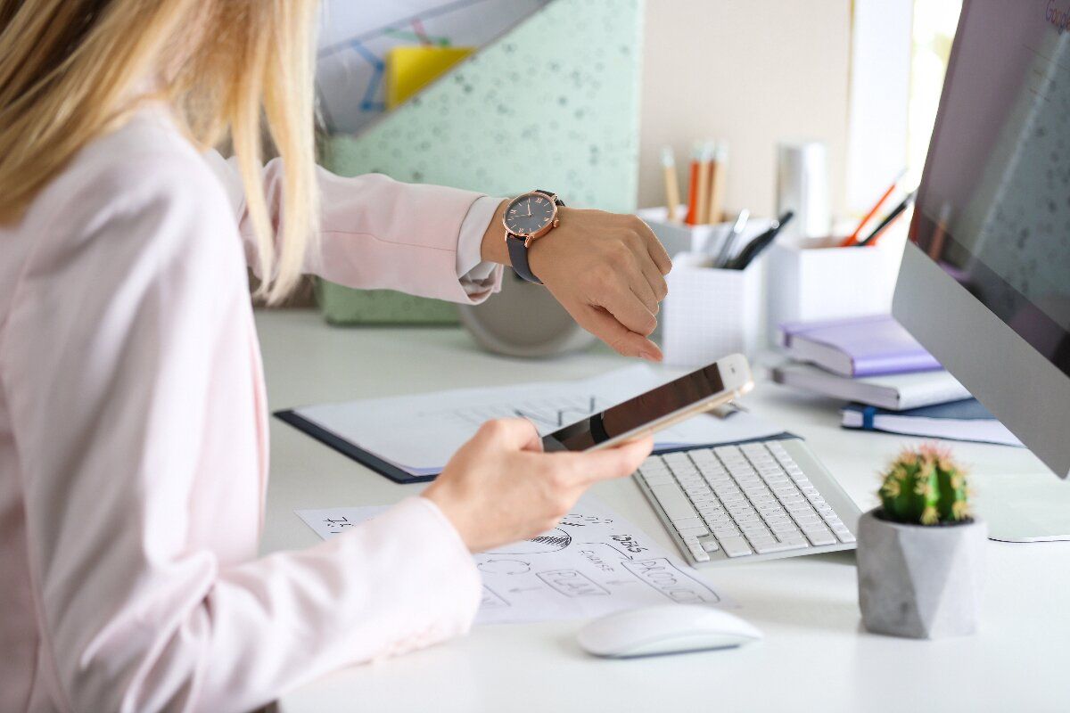 Mitarbeiterin am Schreibtisch sitzend, mit Smartphone in einer Hand, blickt auf ihre Armbanduhr: Zeitmanagement