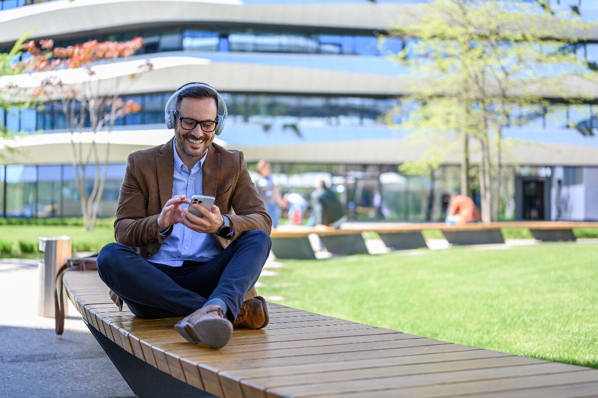 Social Listening: Geschäftsmann mit Kopfhörern sitzt lächeln im Schneidersitz auf einer Bank und blickt auf sein Smartphone