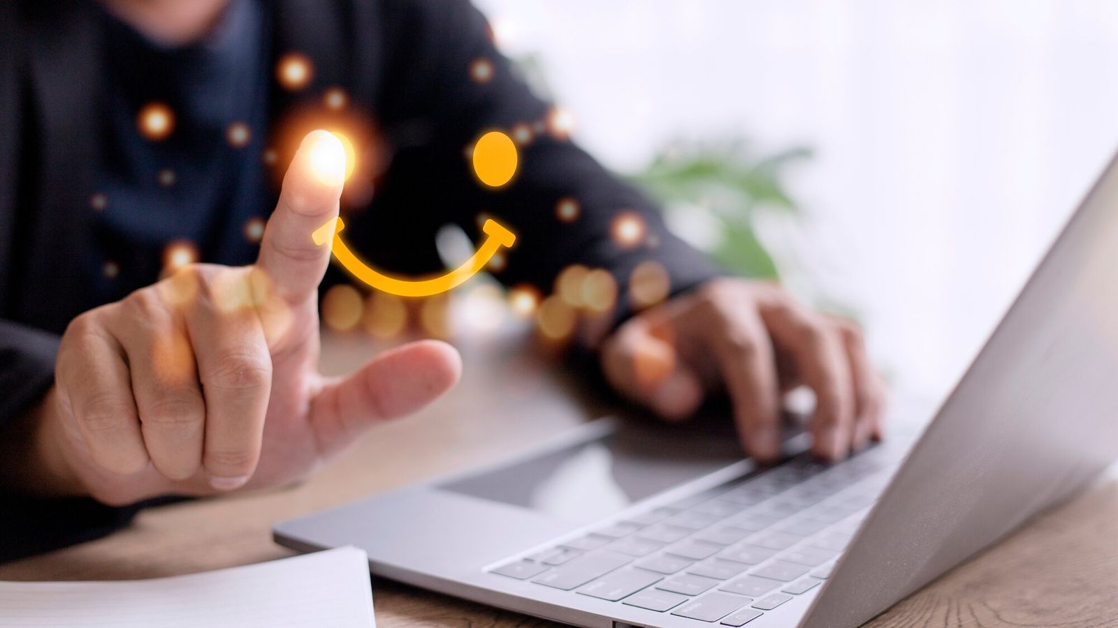 Person sitzt vor einem Laptop und zeichnet mit dem Zeigefinger ein Smiley in die Luft