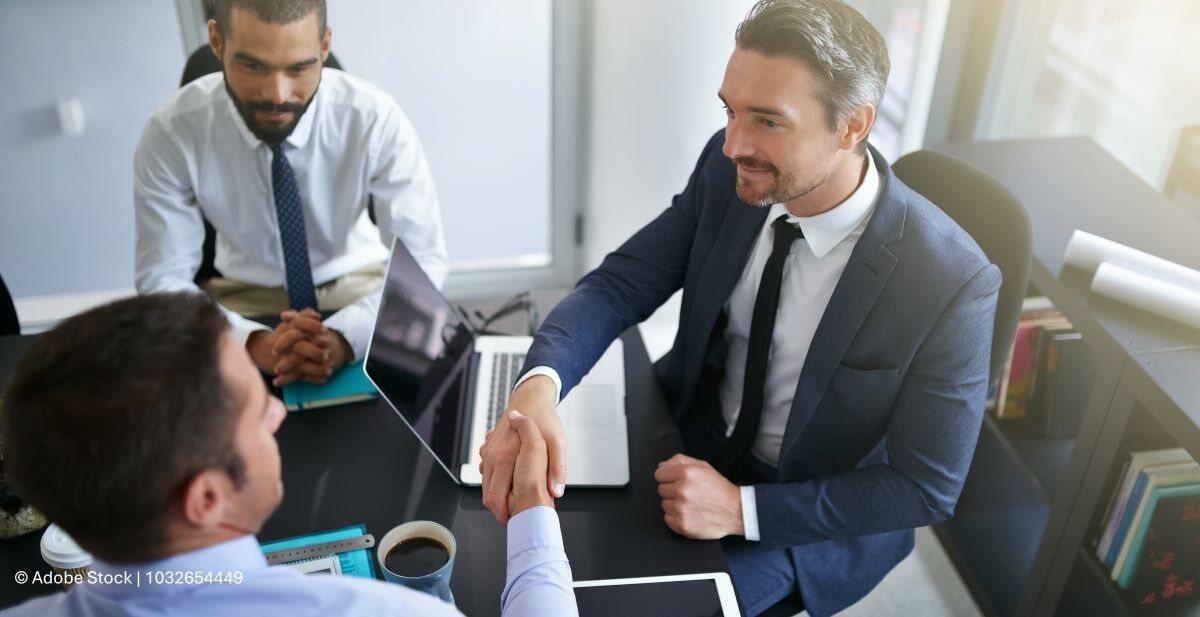 Investition: Zwei Unternehmer schütteln sich die Hände in einem Meetingraum. Im Hintergrund ein dritter Berater.