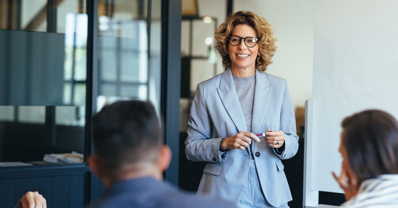 Agile Führung im Change Management:Chefin mittleren Alters in Blazer und Brille präsentiert vor Team.