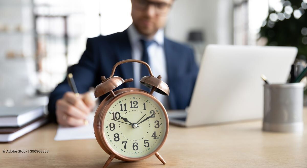 Zeitmanagement: Unternehmer im Arbeitsprozess arbeitet am Laptop und schreibt auf ein Dokument. Großer Wecker im Vordergrund.